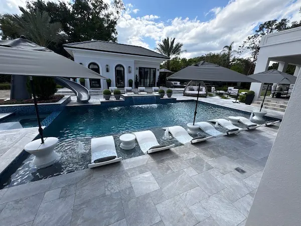 Luxury backyard geometric pool with slate finish on pool decking, row of lounge chairs on a sun shelf, hot tub and waterslide